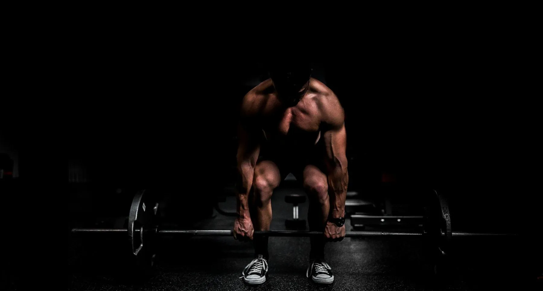 Shirtless lifter crouched over a loaded bar in near-darkness, one hard light on his back